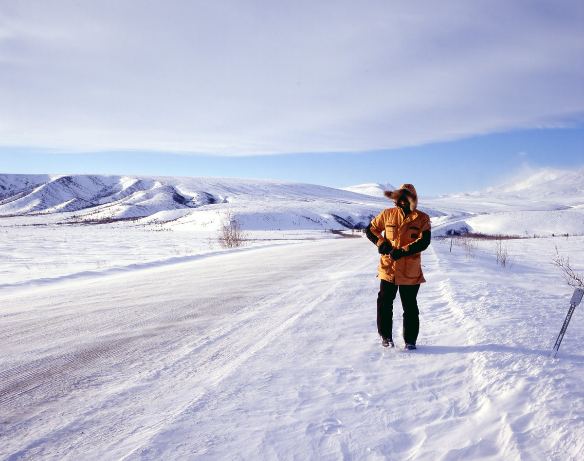 UOMO vestito di un parka giallo con cappuccio che cammina ai margini di una strada ghiacciata in Alaska, spazzata dal vento e circondata da una piana desolata di neve. - Cape Horn