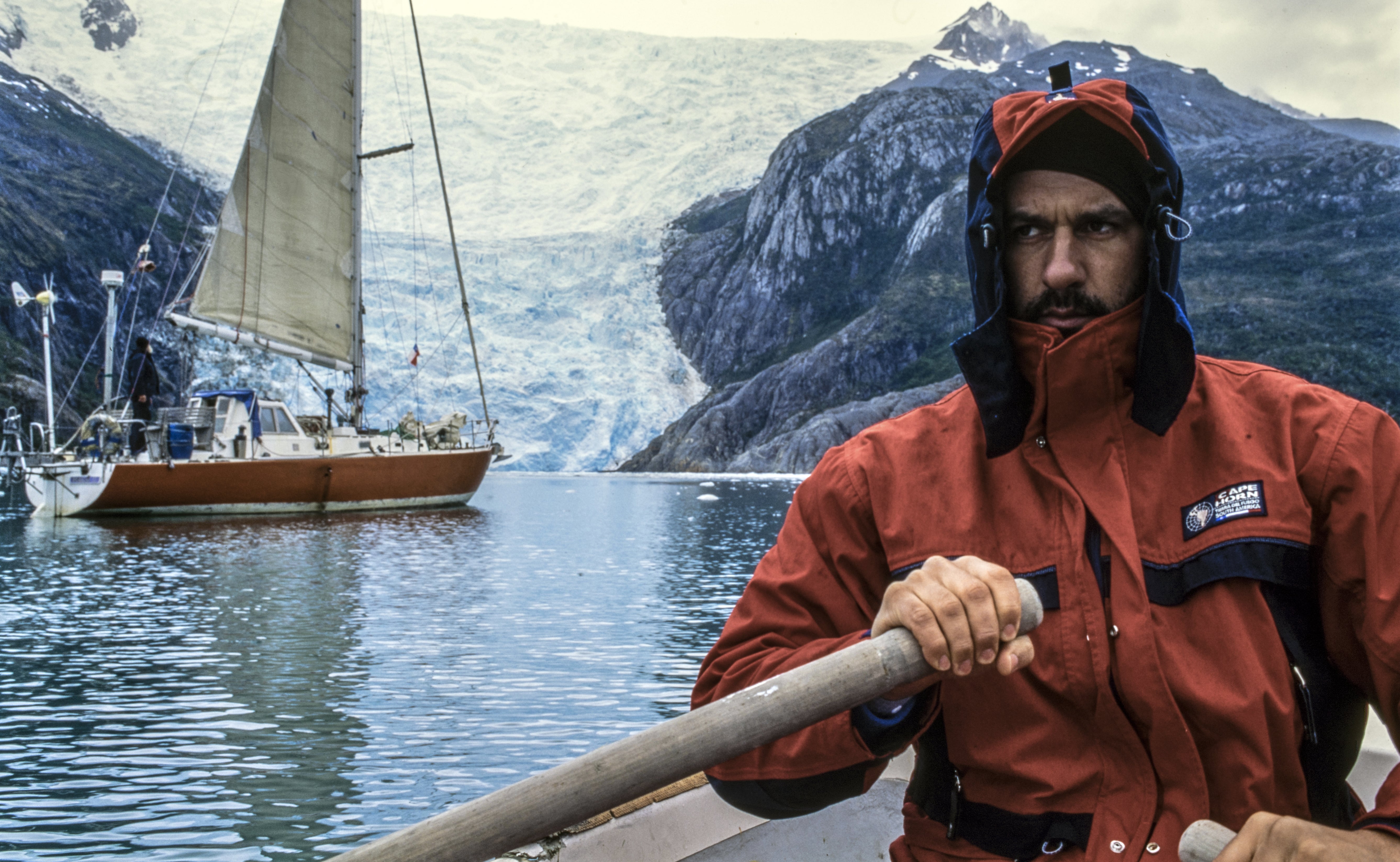 Esploratore con giacca rossa Cape Horn in barca a remi, davanti a un ghiacciaio e a una barca a vela, in un paesaggio estremo. - Cape Horn