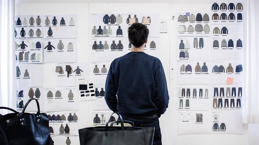 Un designer, visto di spalle, osserva attentamente una parete coperta di bozzetti e schede tecniche di capi d'abbigliamento in un ambiente di studio. L'immagine rappresenta il processo creativo e la cura per il design italiano di Cape Horn, dalla concezione alla realizzazione dei capi - Cape Horn