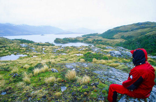 Una persona con abbigliamento tecnico rosso siede osservando un vasto paesaggio naturale con montagne, un lago e vegetazione selvaggia. L'immagine simboleggia l'impegno di Cape Horn verso la natura e la sostenibilità, evocando il rispetto per l'ambiente e l'esplorazione responsabile - Cape Horn