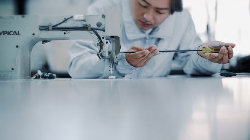 Un artigiano o tecnico si china su una macchina da cucire industriale, concentrato nella regolazione di un meccanismo con un cacciavite. L&#39;immagine simboleggia la cura artigianale, la precisione della manifattura e l&#39;applicazione della tecnologia avanzata nella creazione dei capi Cape Horn - Cape Horn